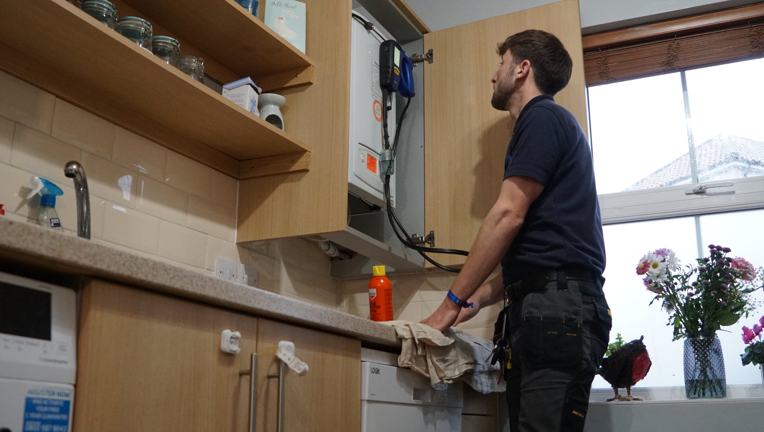 A man working on an electrical panel in a kitchen, cleaning the countertop near a bottle of cleaning spray, with flowers on the window sill.