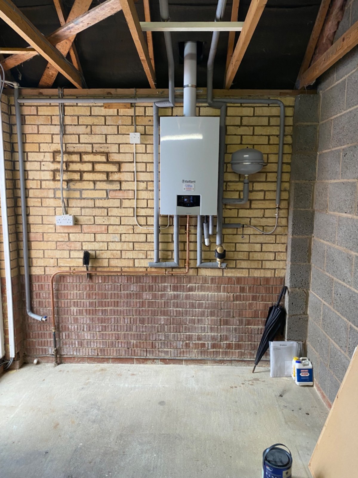 A utility room with a brick wall and exposed wooden ceiling beams. It features a white Vaillant boiler, gray pipes, and a gray expansion tank. There is an electrical outlet, a garden hose, a small brush, and cleaning supplies on the floor.