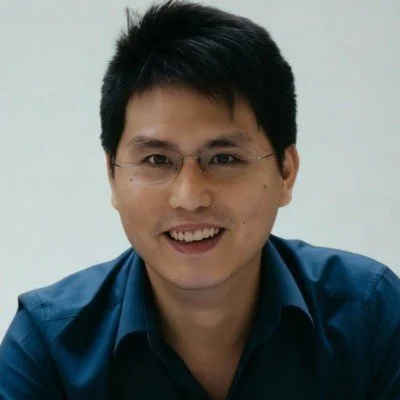 A young man with short black hair and glasses, smiling, wearing a blue collared shirt, against a plain light-colored background.