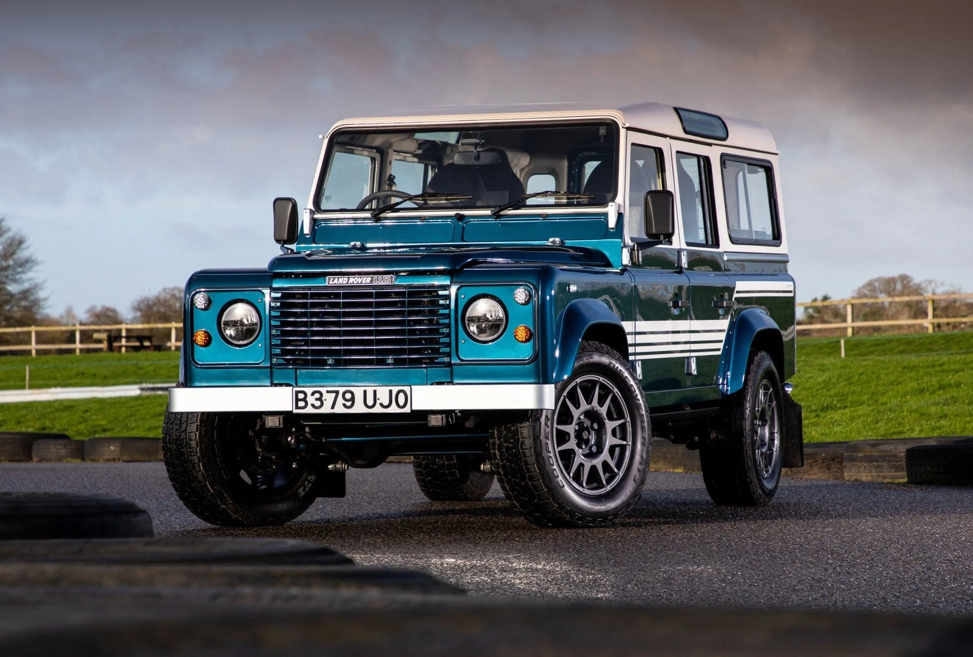 A blue vintage Land Rover SUV parked on a paved surface with a grassy field and cloudy sky in the background.