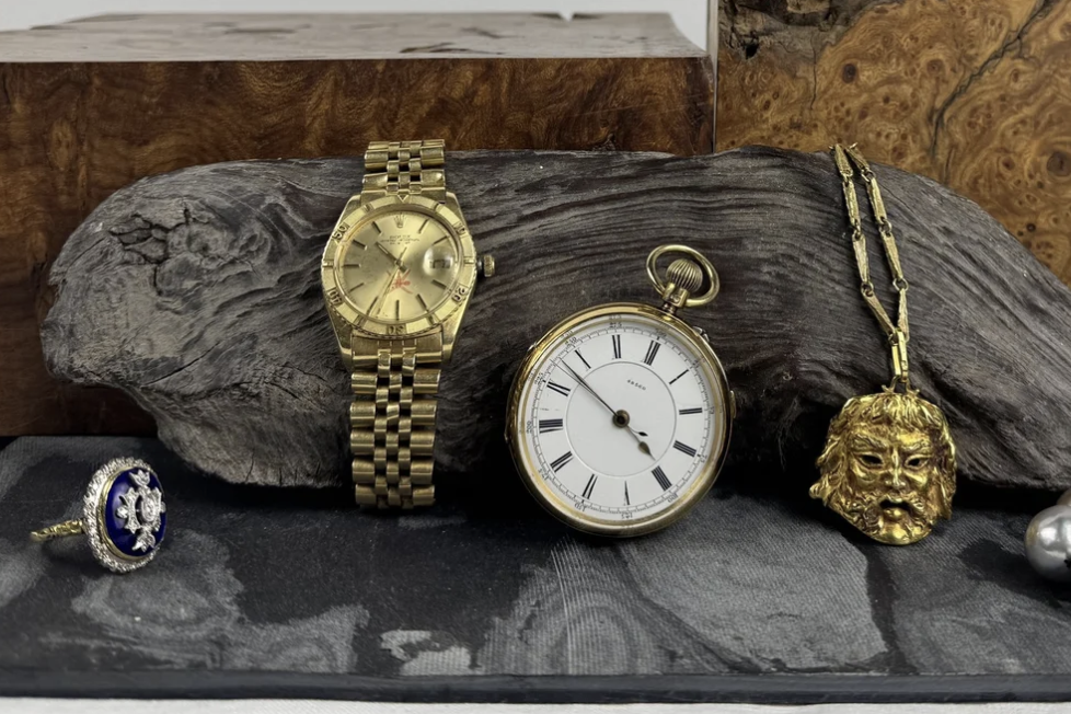 Collection of jewelry and watches displayed on a wooden and fabric surface, including a gold watch, a black and gold pocket watch, a gold lion head necklace, a ring with a blue gemstone, and pearl earrings.