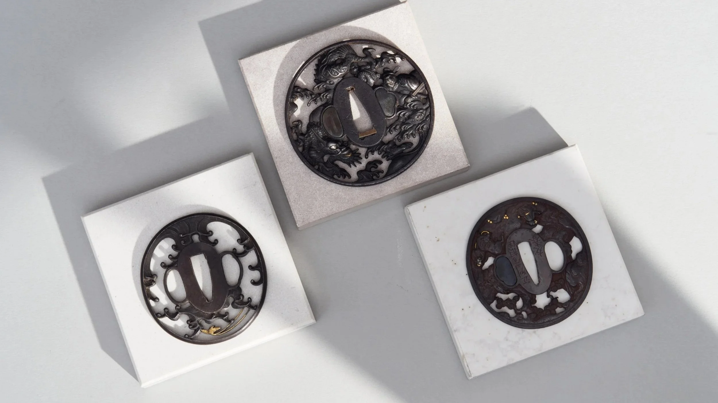 Three Japanese sword guard (menuki) plaques, each with intricate dragon designs, displayed on white and gray square platforms.