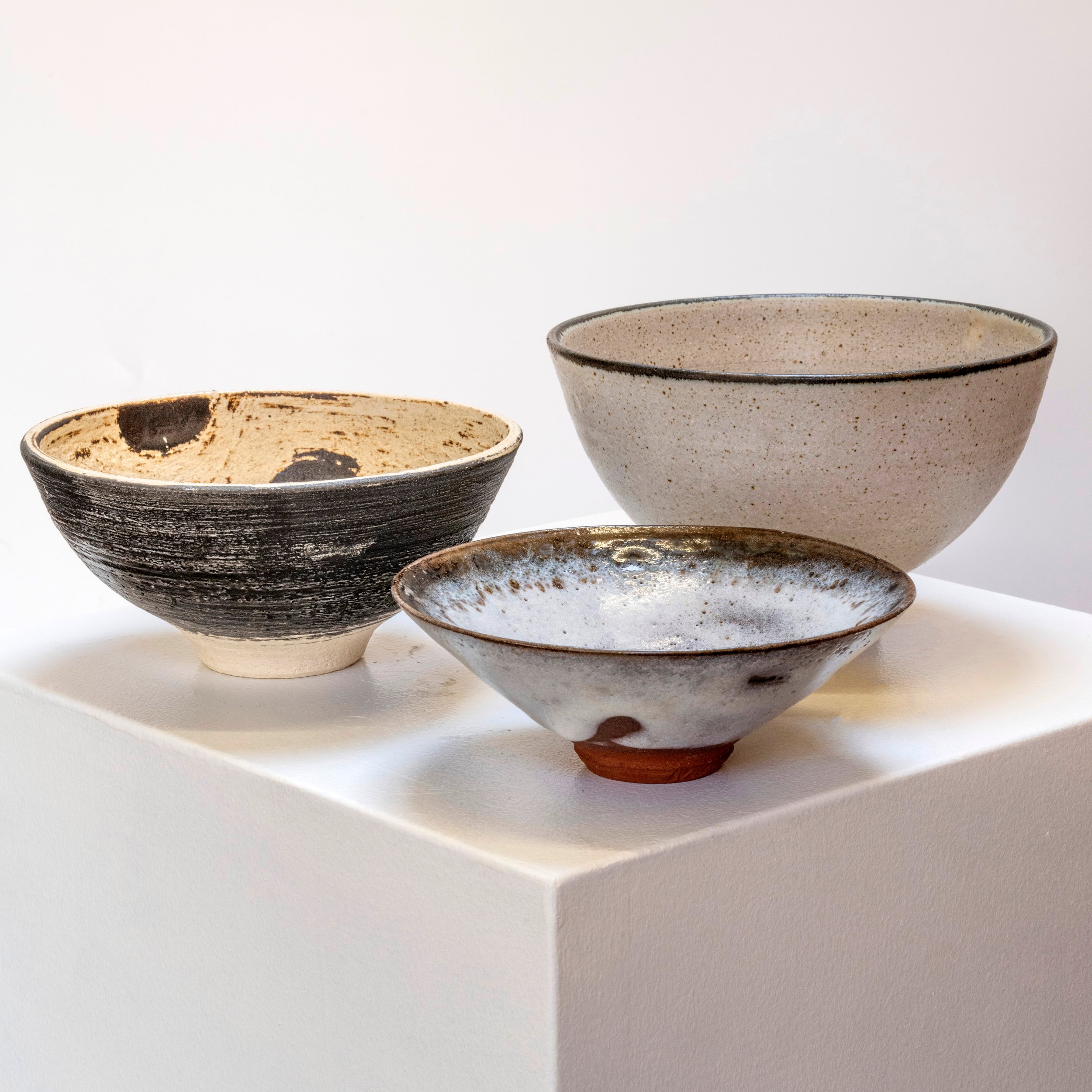 Three ceramic bowls with different glazes and sizes on a white pedestal against a plain background.