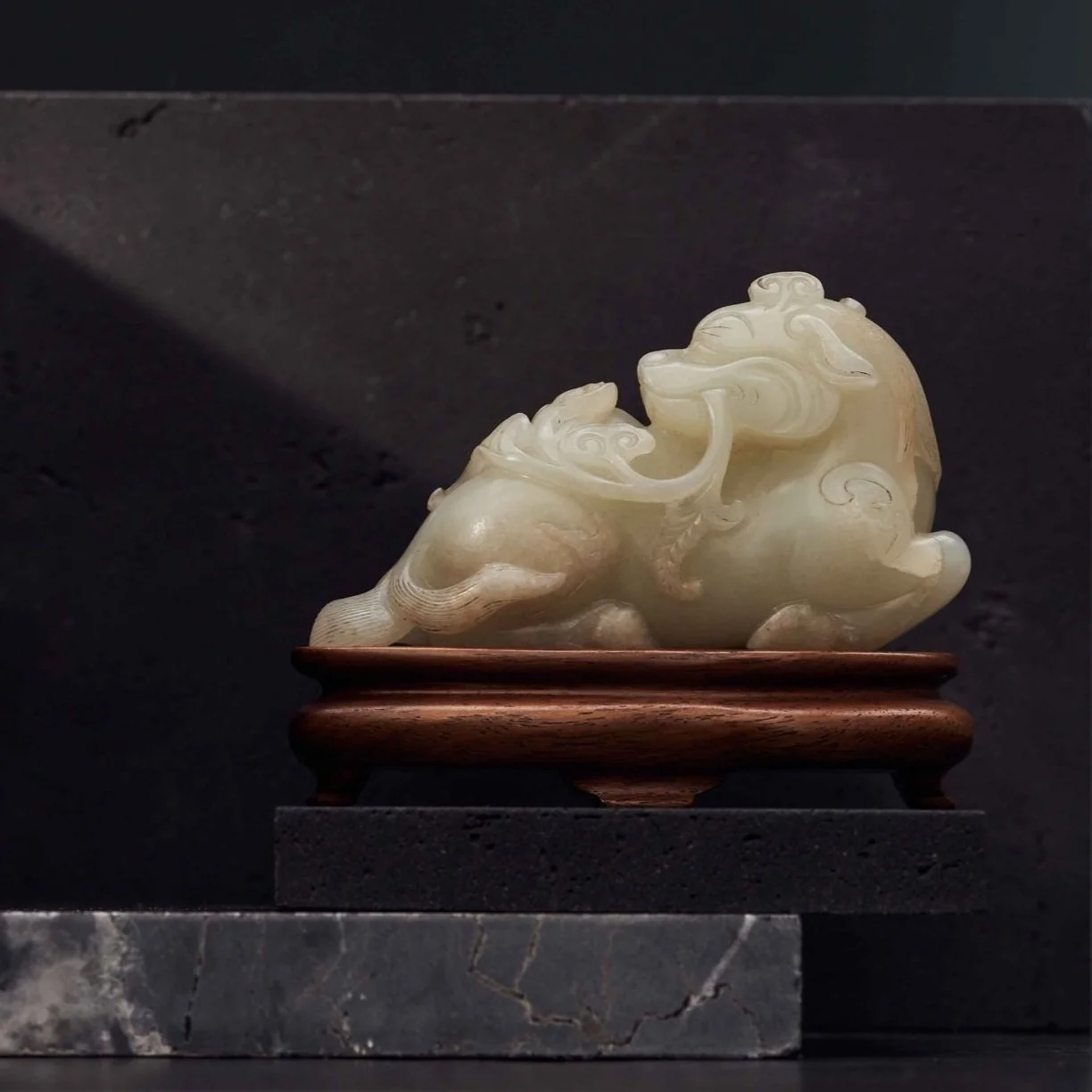A carved jade sculpture of a mythical creature, possibly a qilin or dragon, resting on a wooden stand against a dark background.