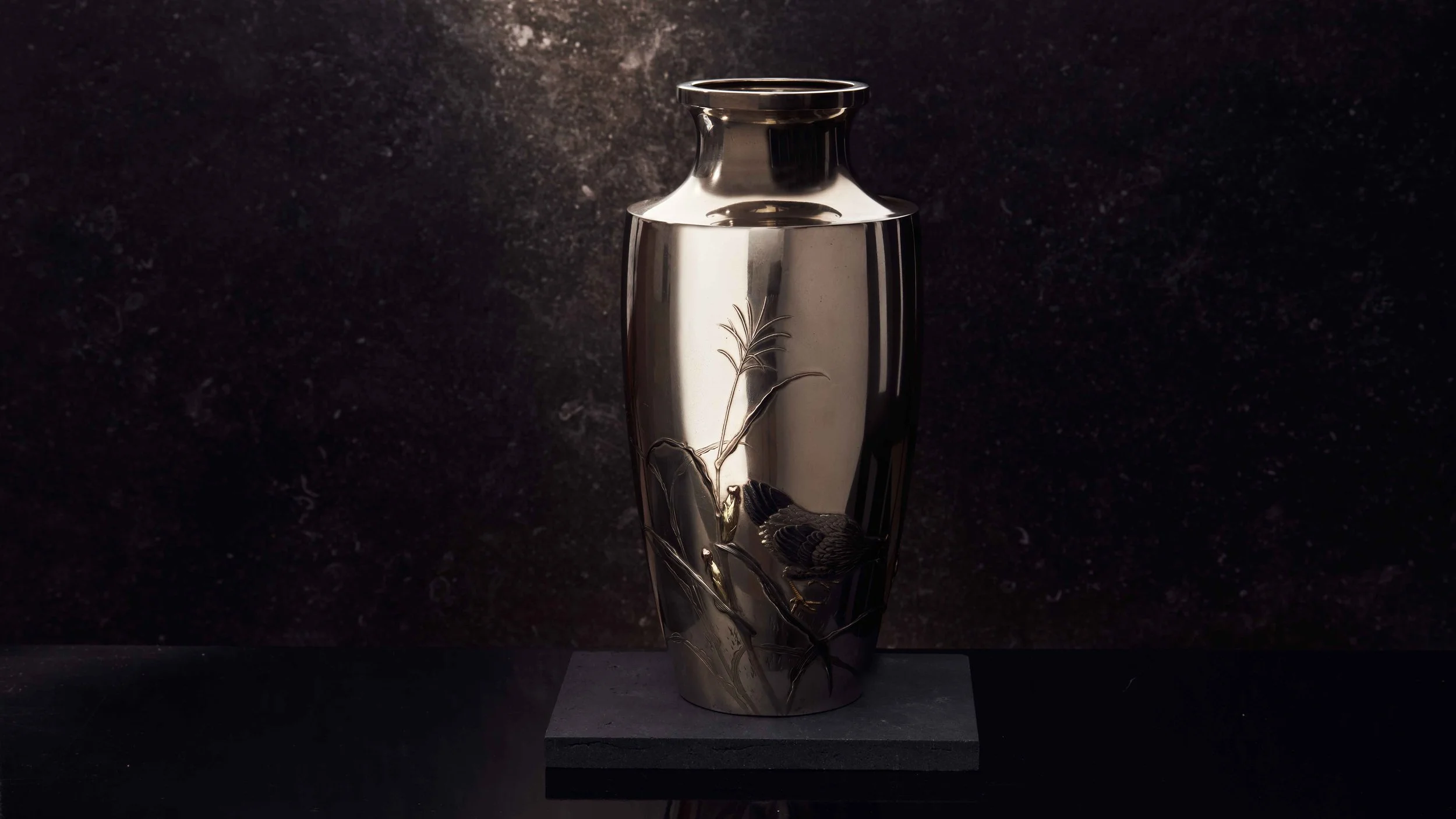 A metallic vase with engraved details of a bird on a branch, placed on a black platform against a dark textured background.
