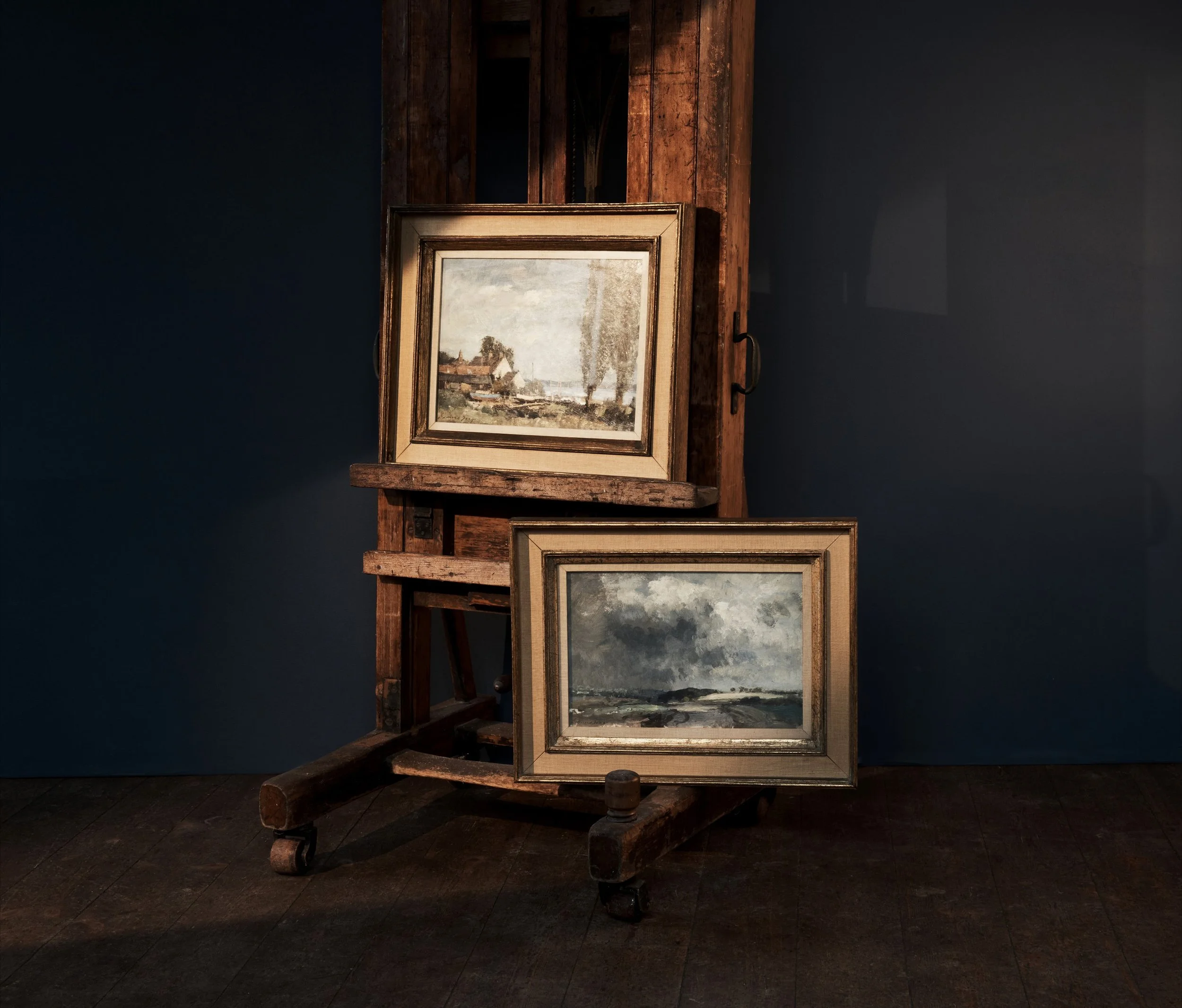 Two landscape paintings leaning against a rustic wooden easel with a dark blue background.