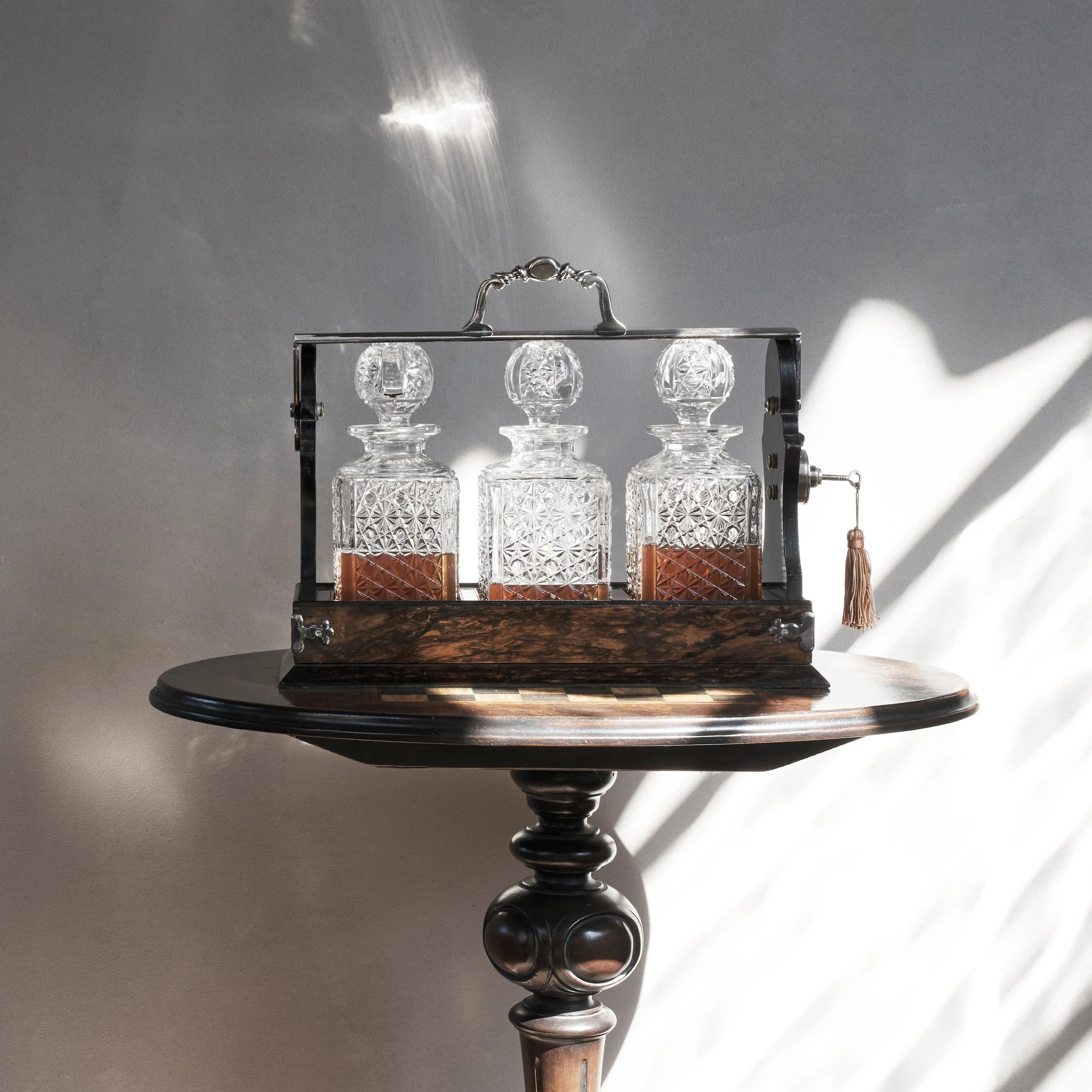 Three glass decanters with diamond-patterned designs, filled with a brown liquid, are displayed in a metal and wood carrier on a round wooden table, with sunlight creating shadows on a plain grey wall.