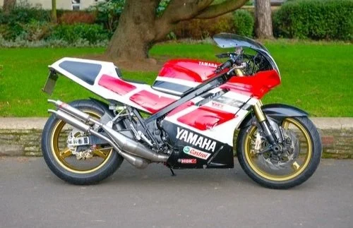 Red and white Yamaha racing motorcycle parked on street with grass and trees in background.