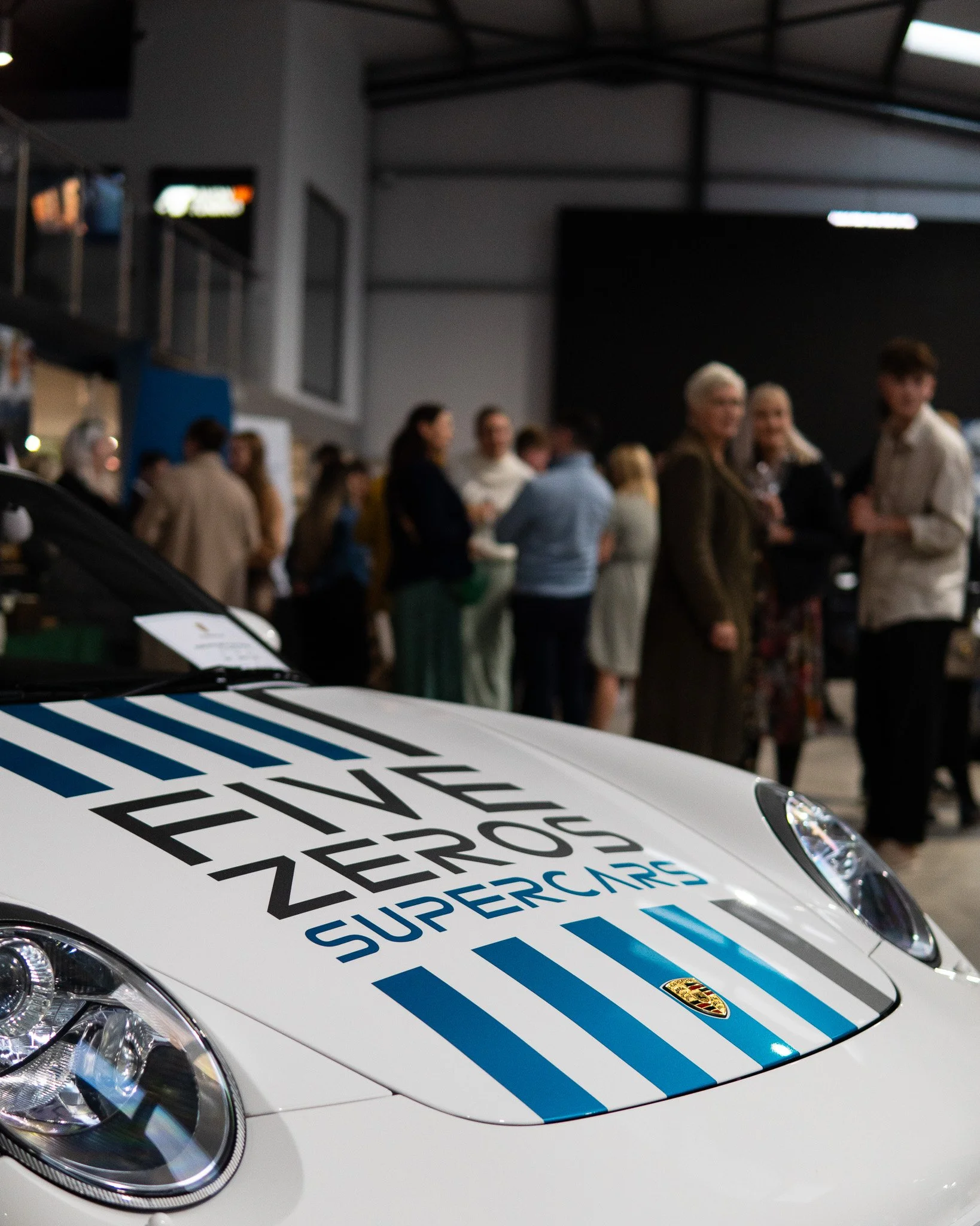 Close-up of the hood of a white Porsche with the British police text 'METROPOLITAN POLICE ZERO EMISSIONS' and blue stripes, with a crowd of people in the background at an indoor event.