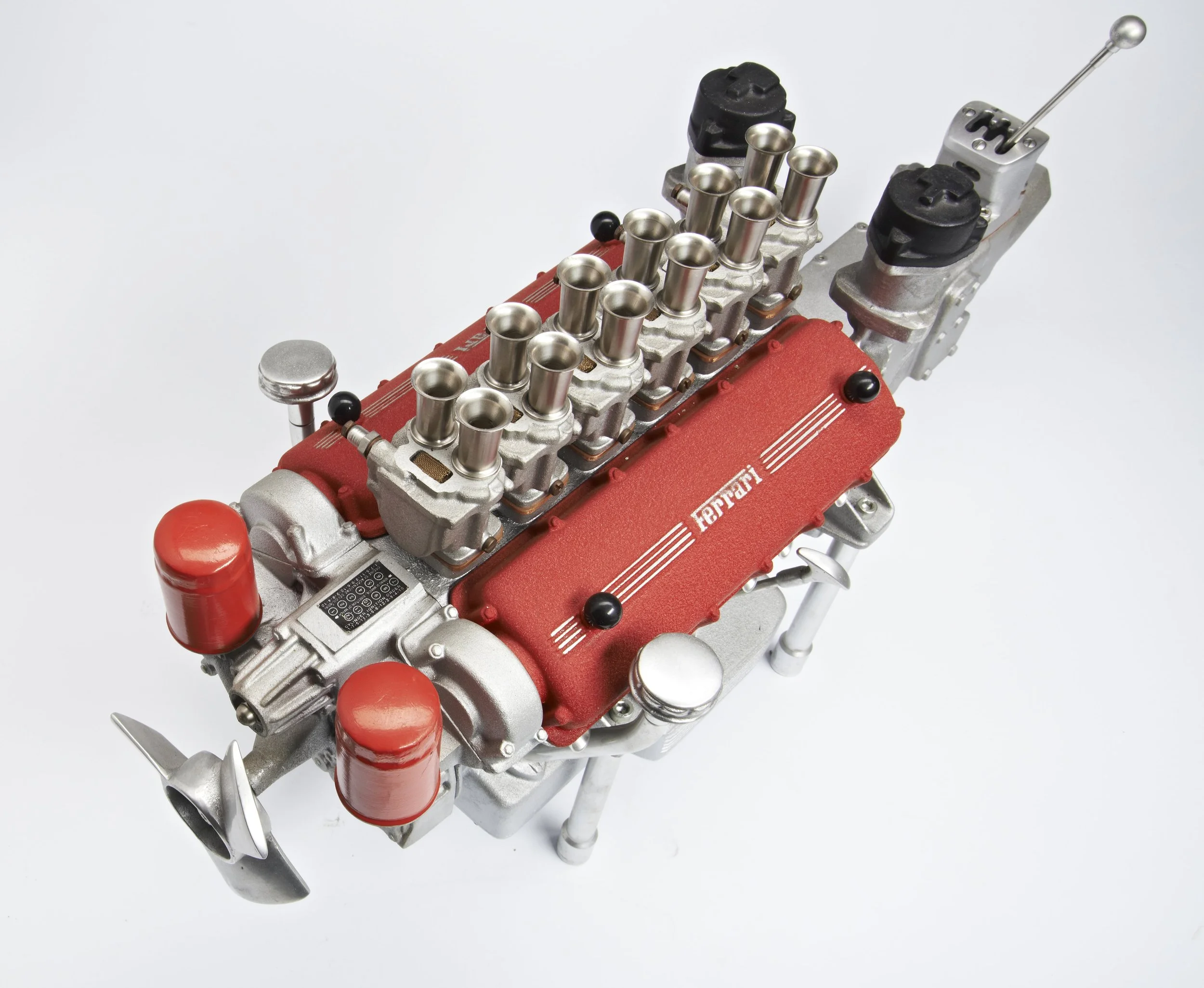 Miniature Ferrari engine model with red valve covers and silver components placed on a white background.