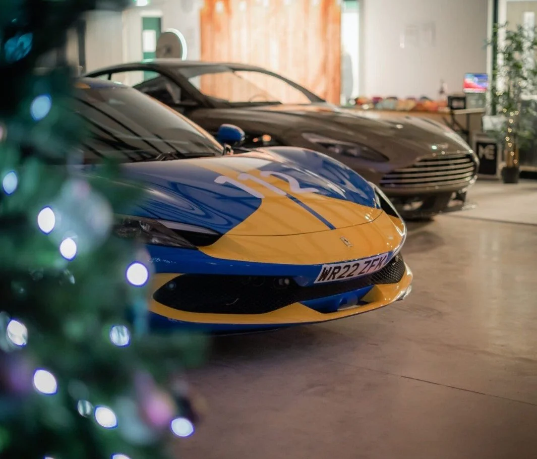 Two luxury sports cars, a blue and yellow car in the front, and a black car behind, are parked indoors near a wooden wall, with some Christmas decorations partly visible on the left.