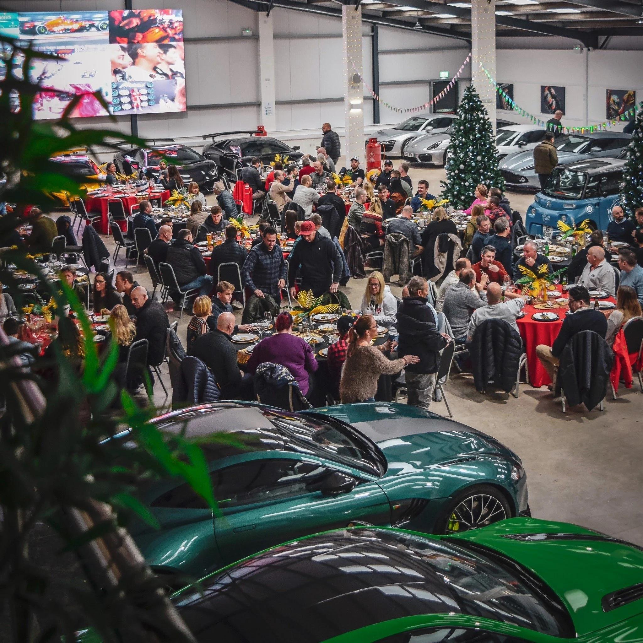 A large gathering of people at an indoor event, with tables decorated with yellow flowers, Christmas trees, and festive decorations. The area is surrounded by luxury cars and includes a large screen displaying racing images.
