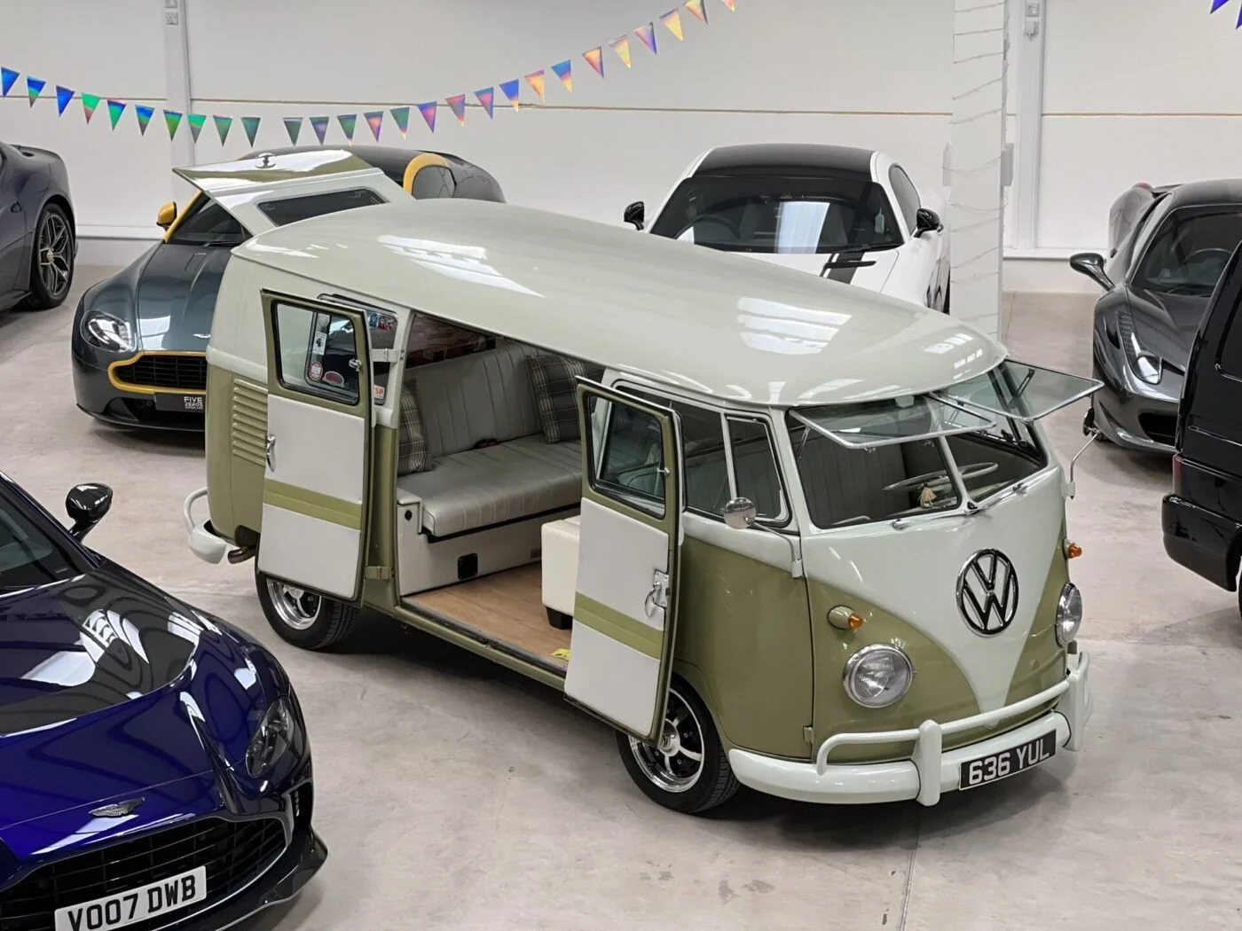 A vintage Volkswagen camper van with doors open, showing an interior with a bench seat, parked among modern luxury cars in an indoor showroom decorated with colorful pennant banners.