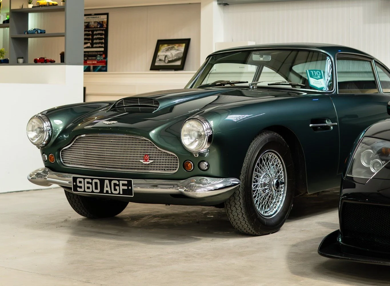 A classic dark green Aston Martin vintage car with wire wheels, parked indoors alongside other luxury vehicles, within a showroom or garage setting.