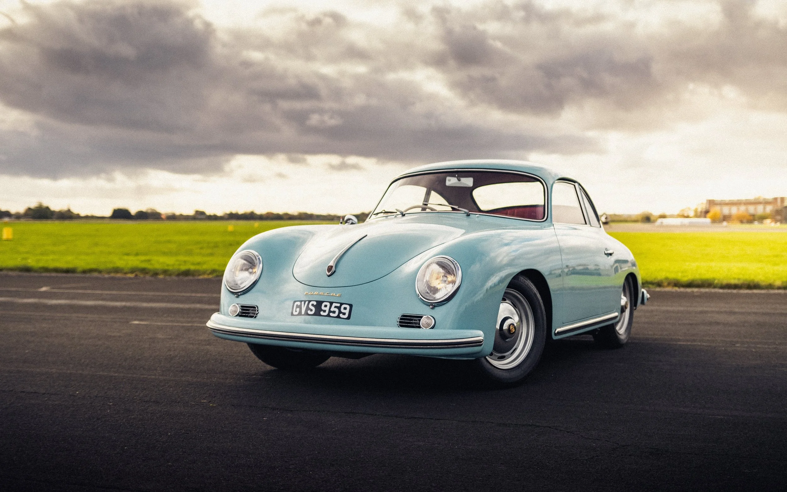 A vintage light blue Porsche 356 parked on a paved surface with a grassy field and cloudy sky in the background.