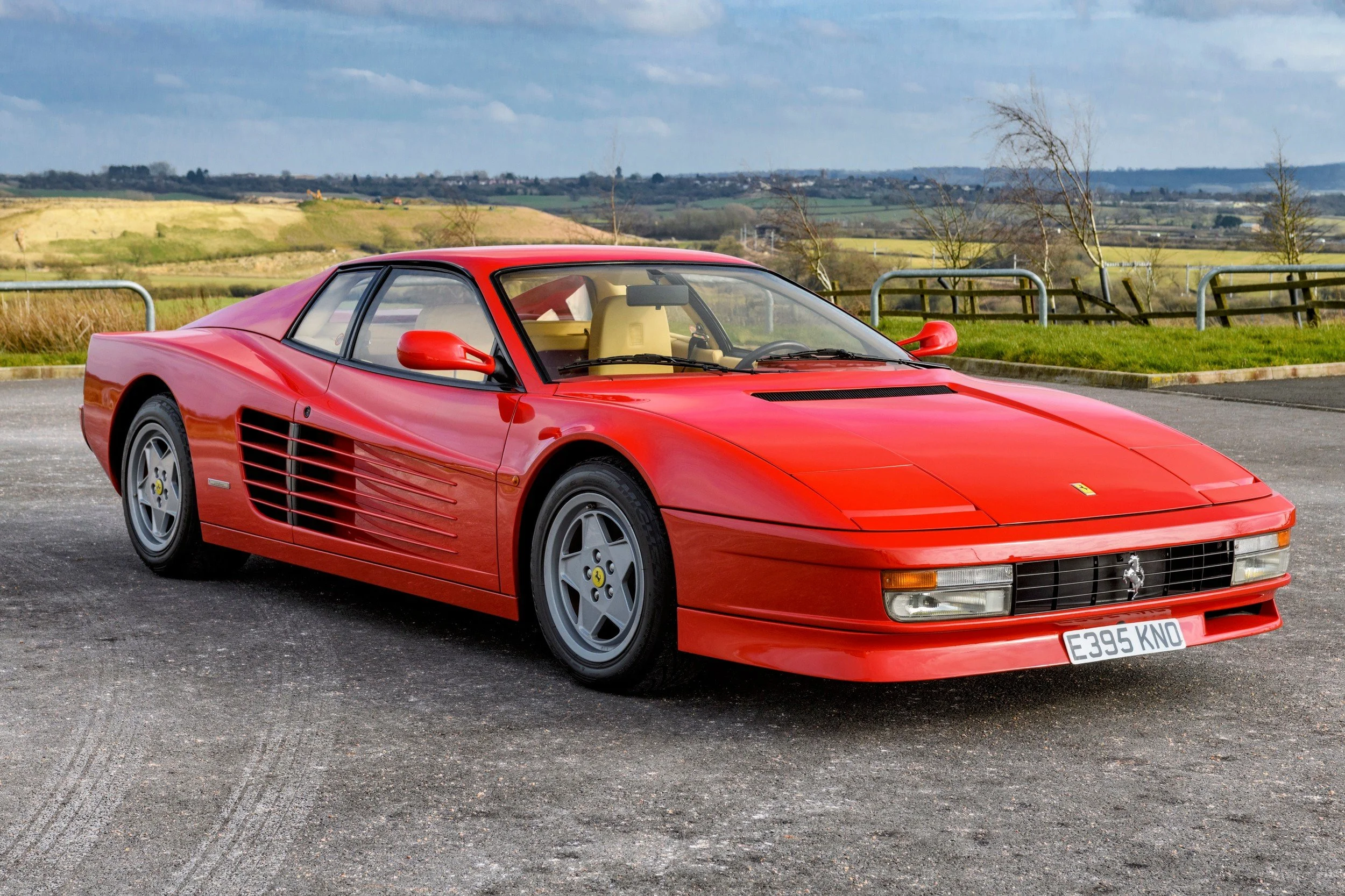 Red Ferrari Testarossa sports car parked on a paved lot with a countryside background.