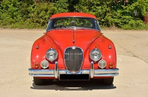 Front view of a vintage red car parked on a sandy surface with green trees in the background.