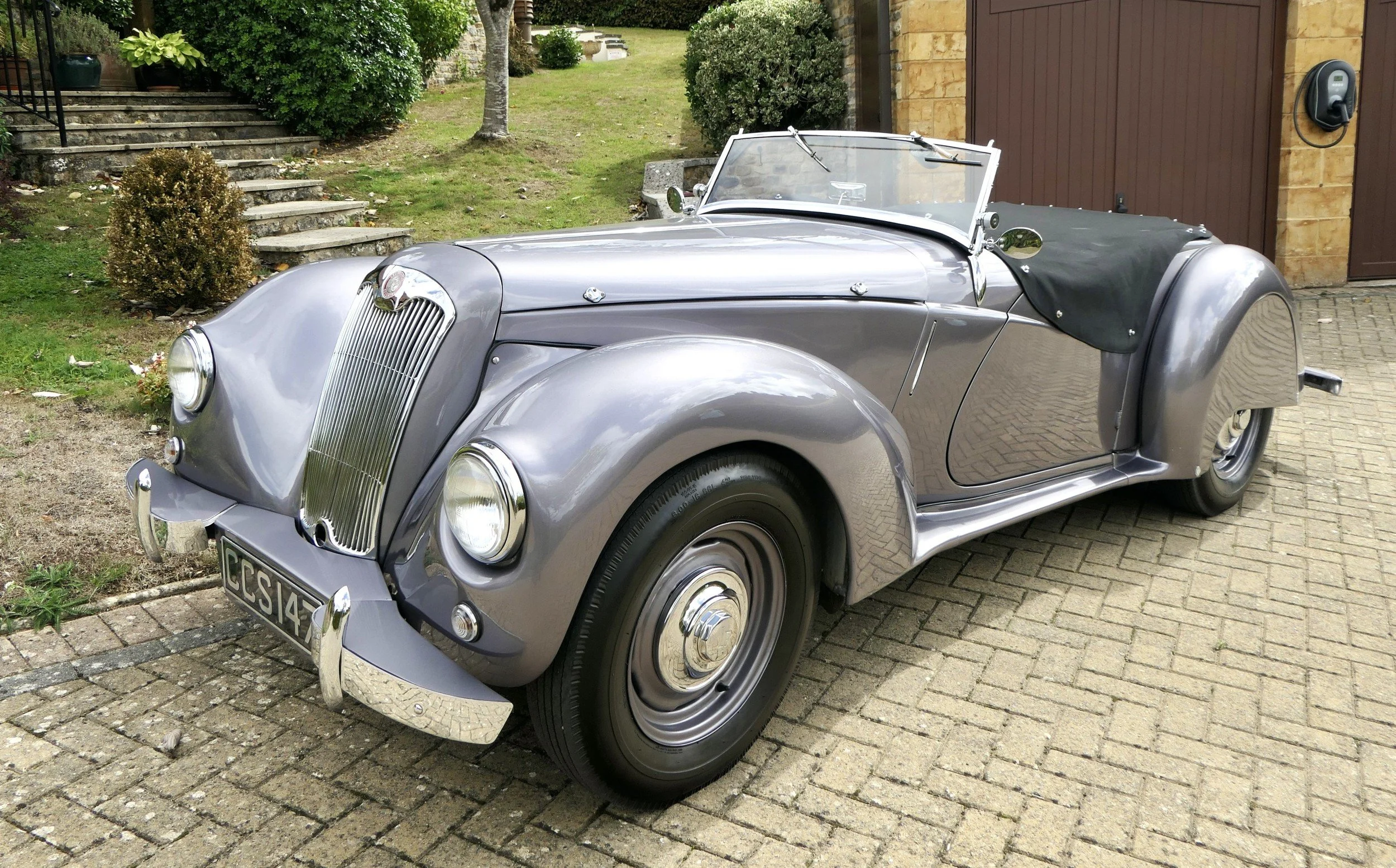 A vintage silver convertible car parked on a brick driveway in front of a house with a wooden garage door and a brown wall.