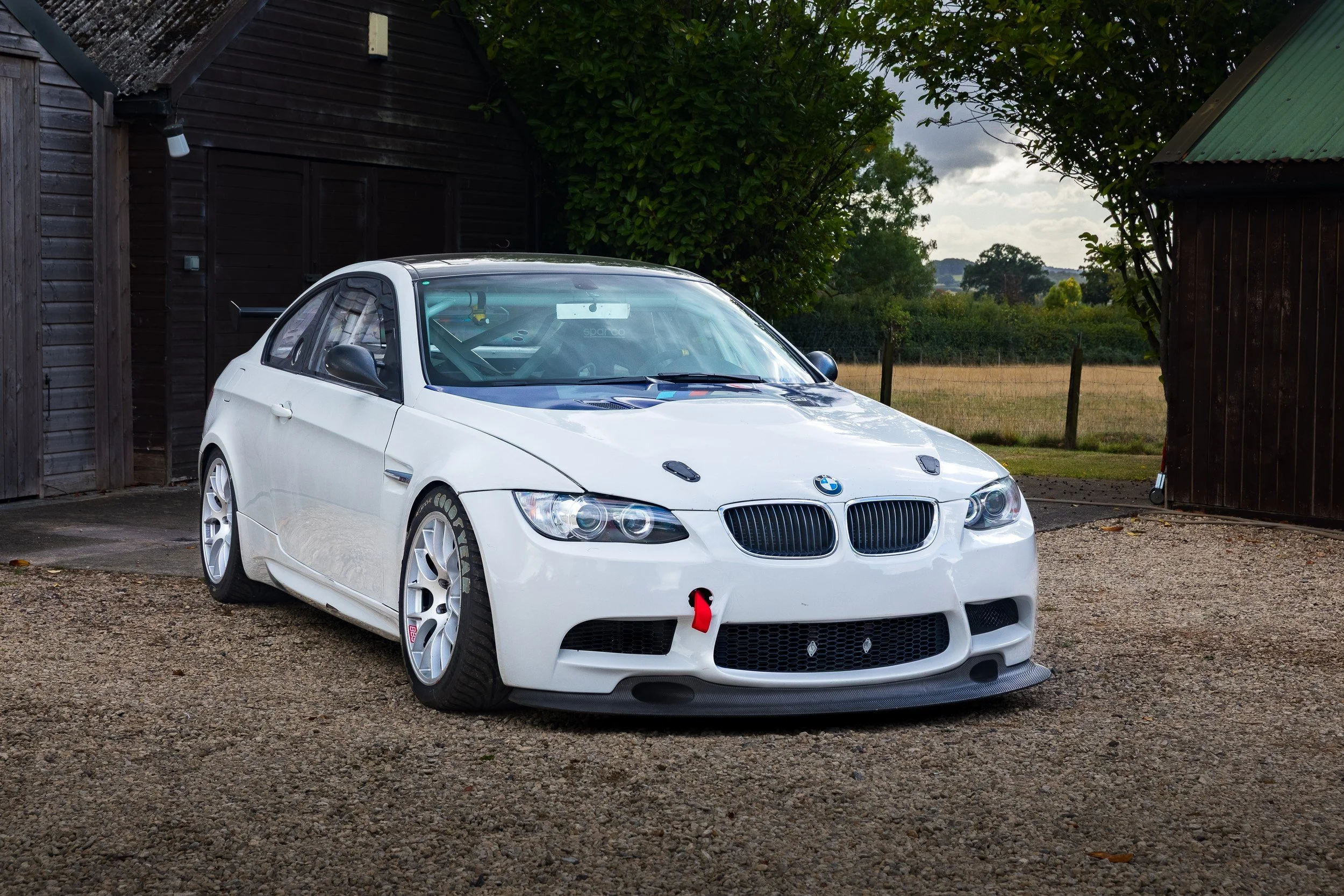 A white BMW race car parked on a gravel driveway, with a tow hook on the front and a racing harness visible inside, surrounded by a rural setting with trees and farm buildings.
