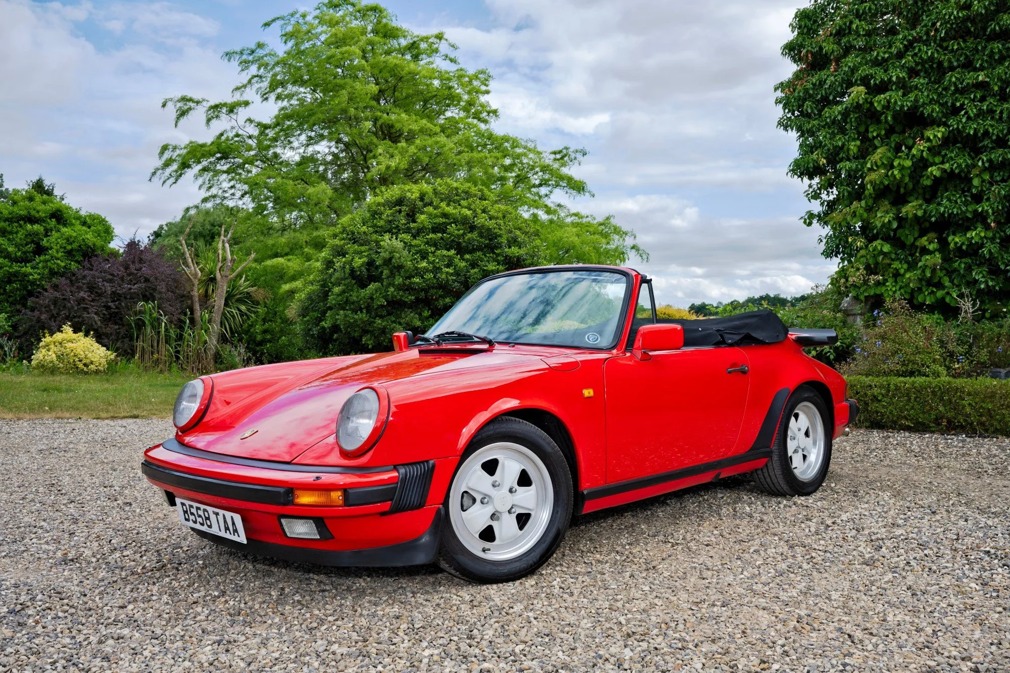 Red classic convertible sports car parked on gravel ground, with green trees and cloudy sky in the background.