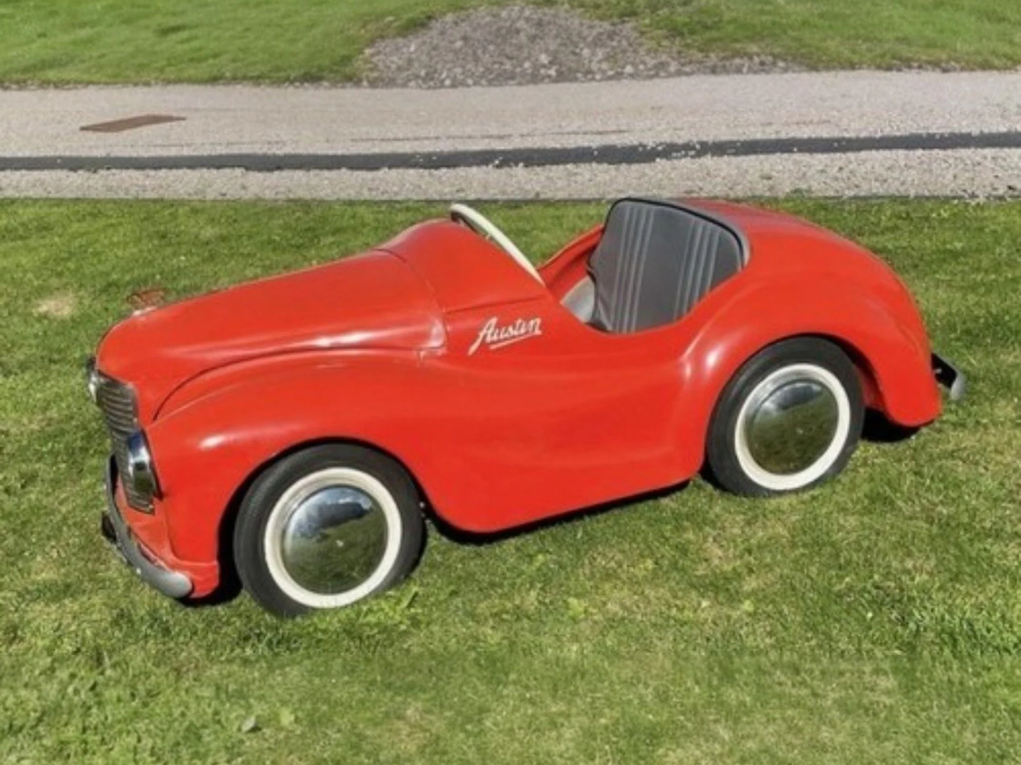 A red vintage pedal car designed to look like a classic automobile, parked on a grassy area near a sidewalk.