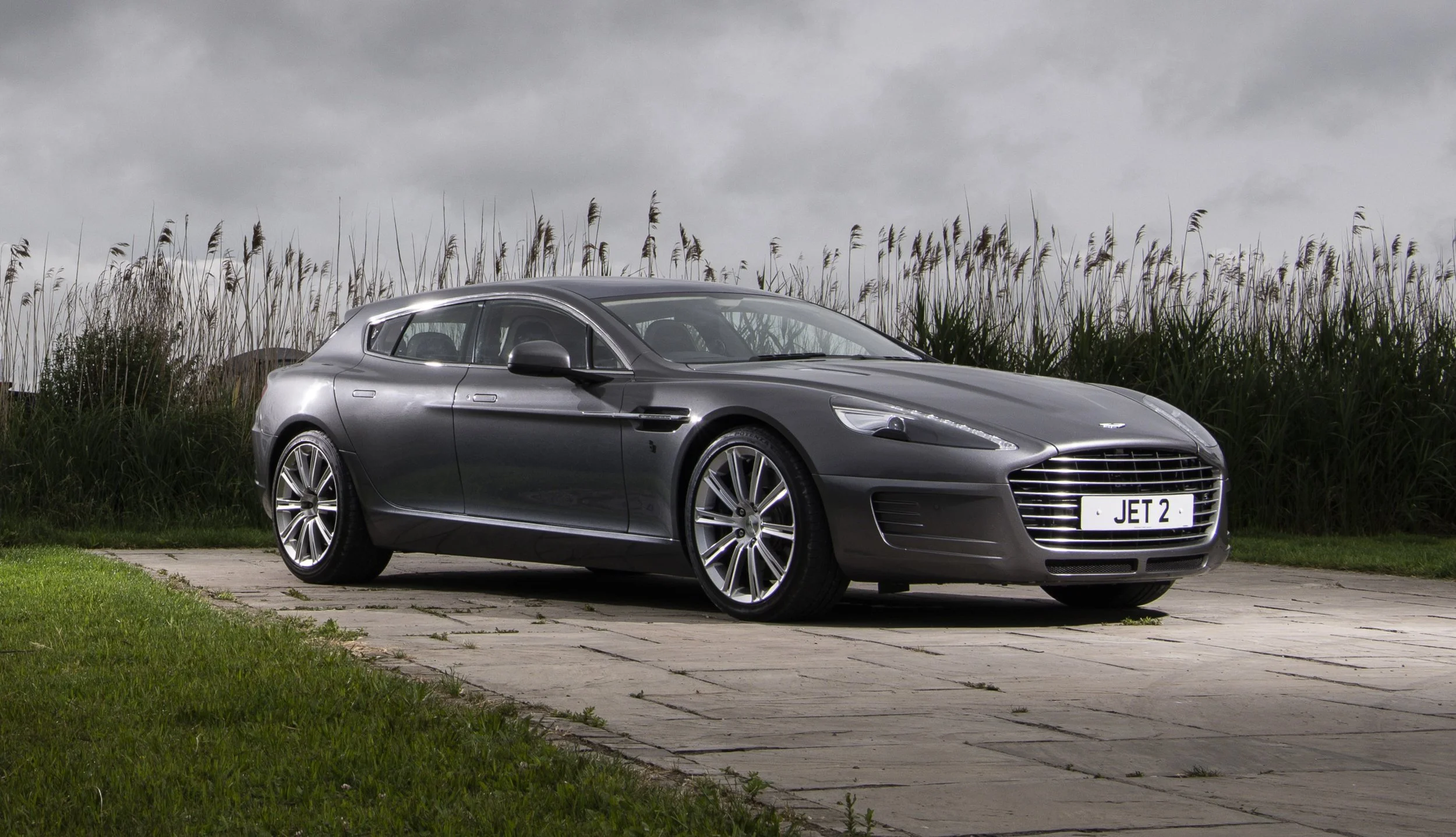 A sleek, gray Aston Martin sports car parked on a paved driveway in front of tall grass against a cloudy sky.