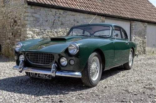 Green vintage car parked on gravel in front of a stone building with a tiled roof.