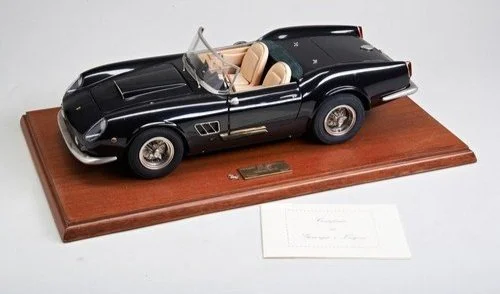 A detailed model of a black vintage convertible car on a wooden display base with a small plaque and a sheet of paper nearby.