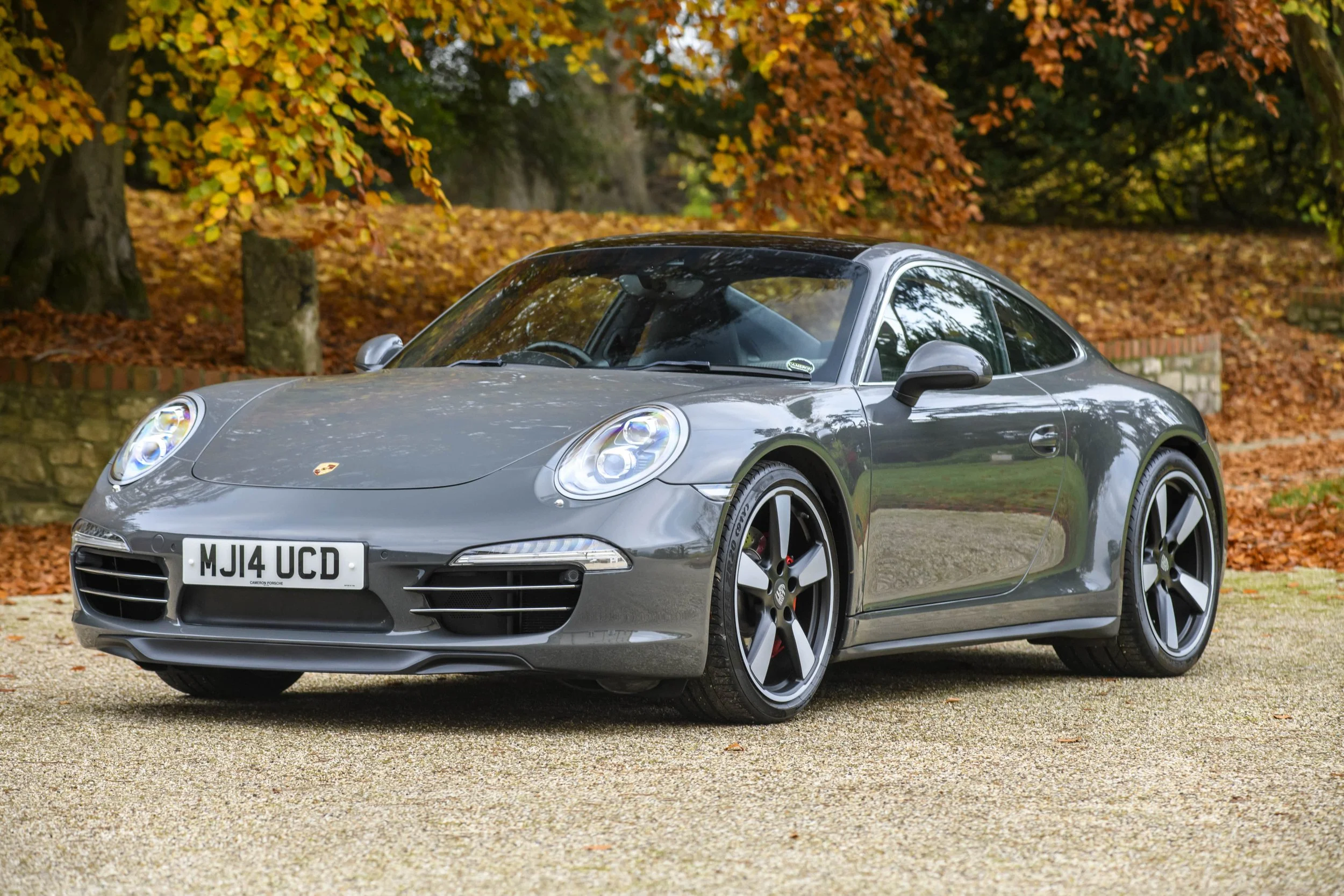 A gray Porsche sports car parked on a gravel surface with autumn trees and fallen leaves in the background.