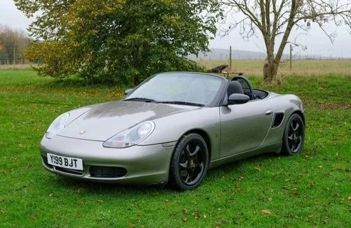 Silver convertible sports car parked on grass with trees and open field in background.