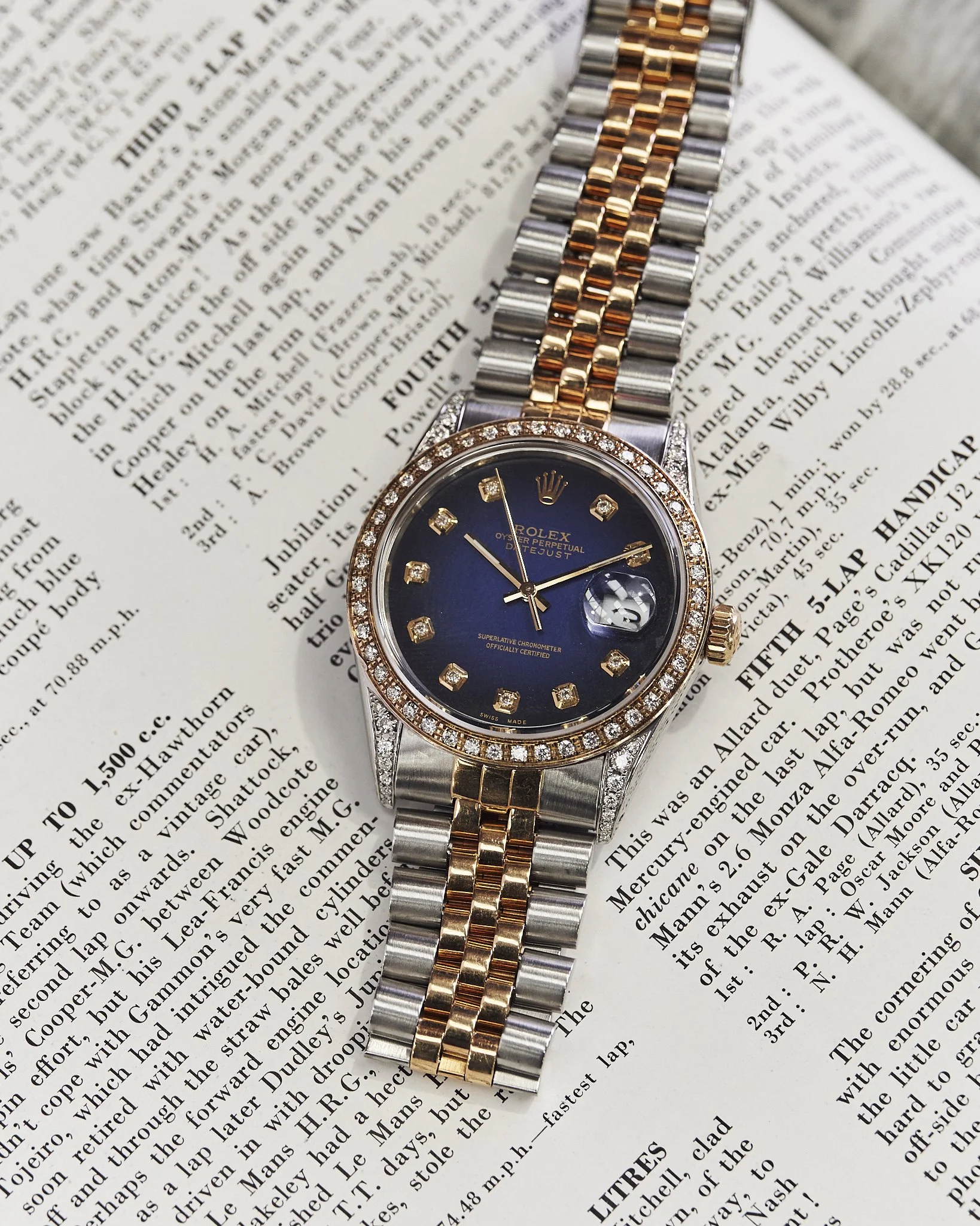 A Rolex Oyster Perpetual Datejust wristwatch with a blue dial, diamond hour markers, a diamond-studded bezel, and a two-tone metal band, placed on an open newspaper.
