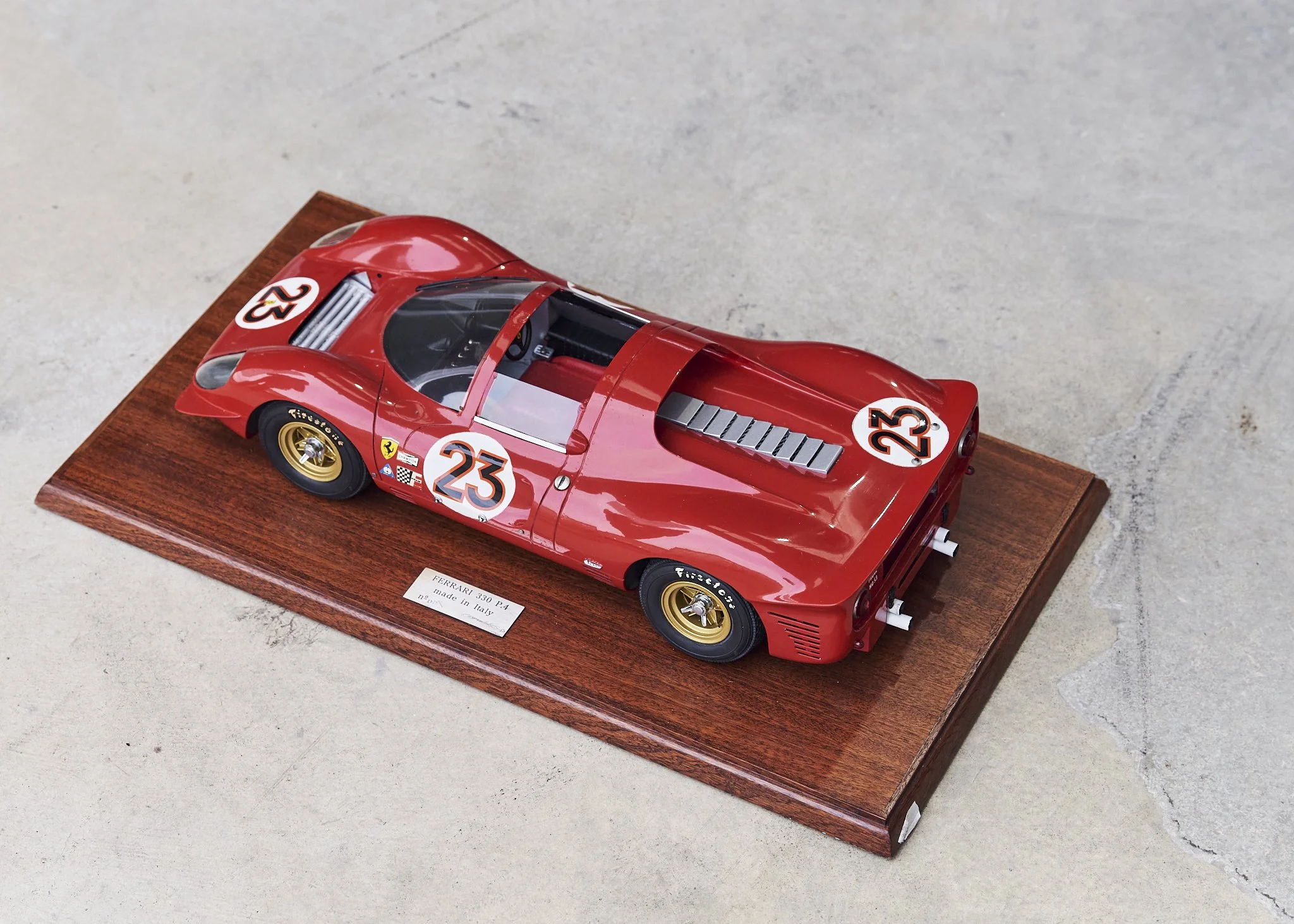 A detailed model of a red Ferrari 330 P4 race car with the number 23 on it, mounted on a wooden display base.