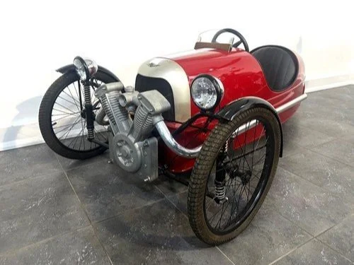 A small red and black vintage-style three-wheeled motorcycle or pedal car with a single seat and handlebars, designed to resemble a racing motorcycle.