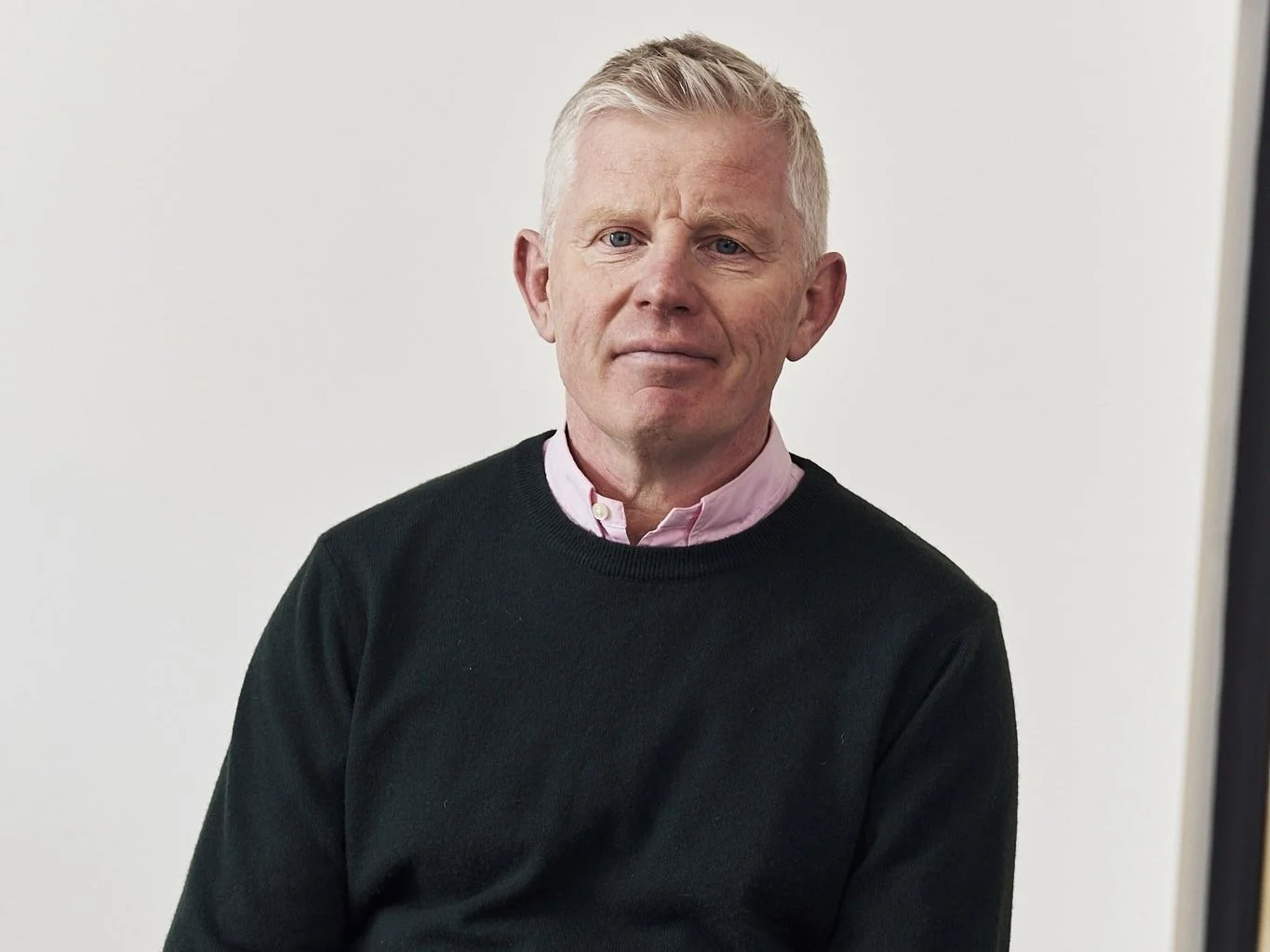A man with blonde hair and blue eyes wearing a black sweater over a pink collared shirt against a plain white background.