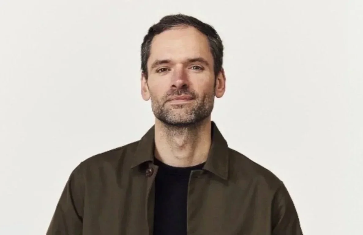 A man with short dark hair and a beard wearing a dark green shirt, standing against a plain white background.