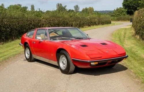 A red vintage sports car parked on a gravel path with green bushes and trees in the background.
