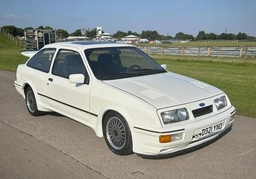 A white vintage Ford Sierra hatchback car parked on a paved road near a grassy field and racetrack in the background.