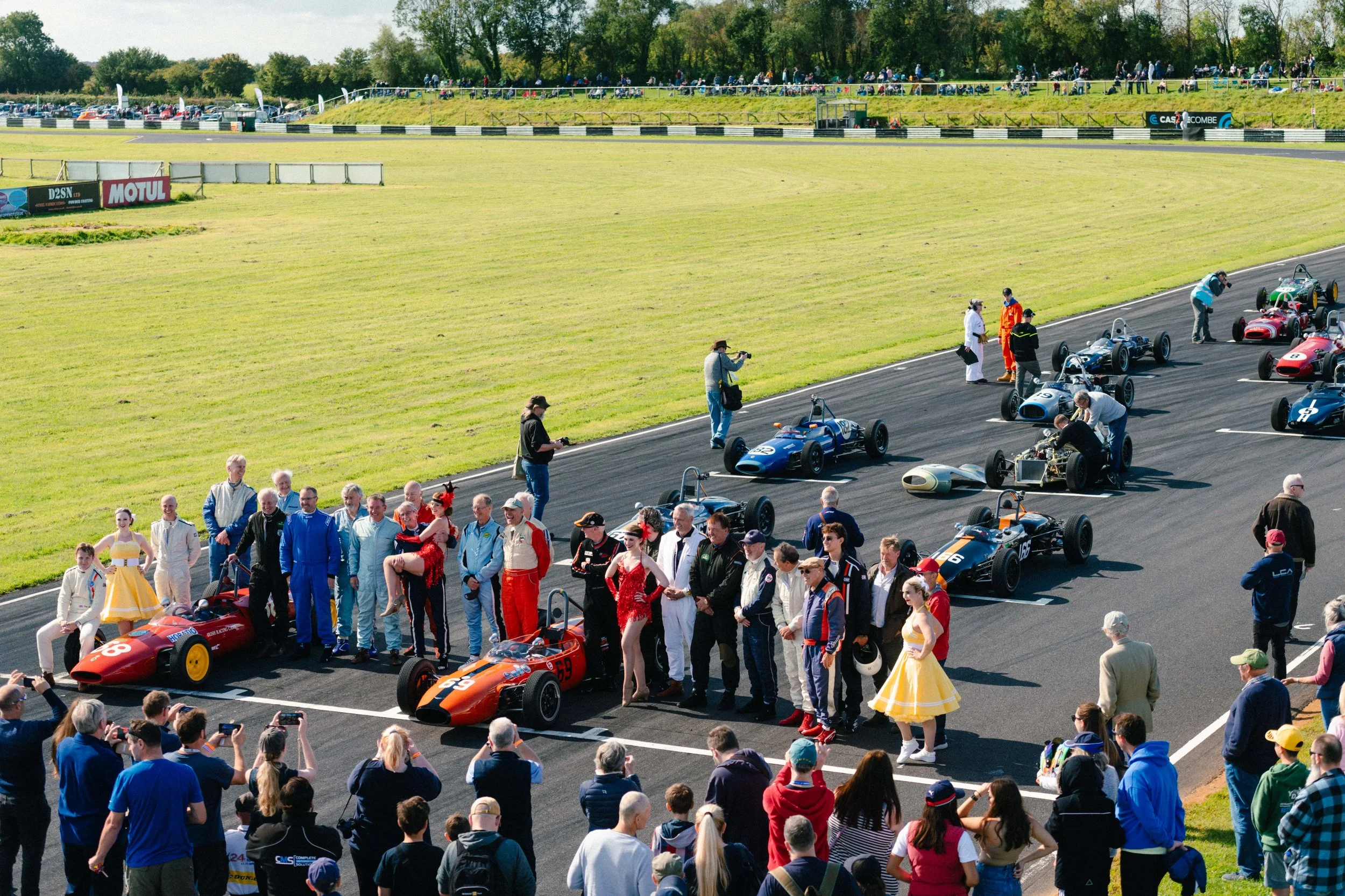 Vintage race cars lined up on a racetrack with drivers, crew, and spectators gathered around; some people taking photos, others conversing. The scene is set on a sunny day with a grassy field and trees in the background.