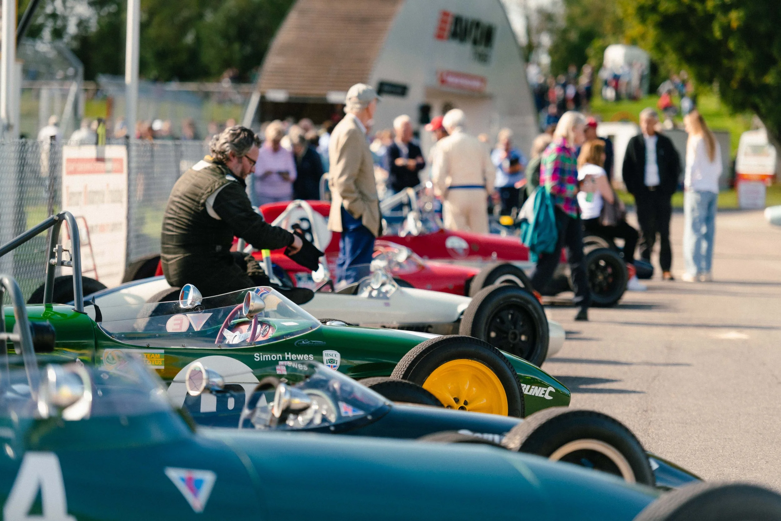 Classic race cars parked in a row at an outdoor event with people walking and chatting in the background.