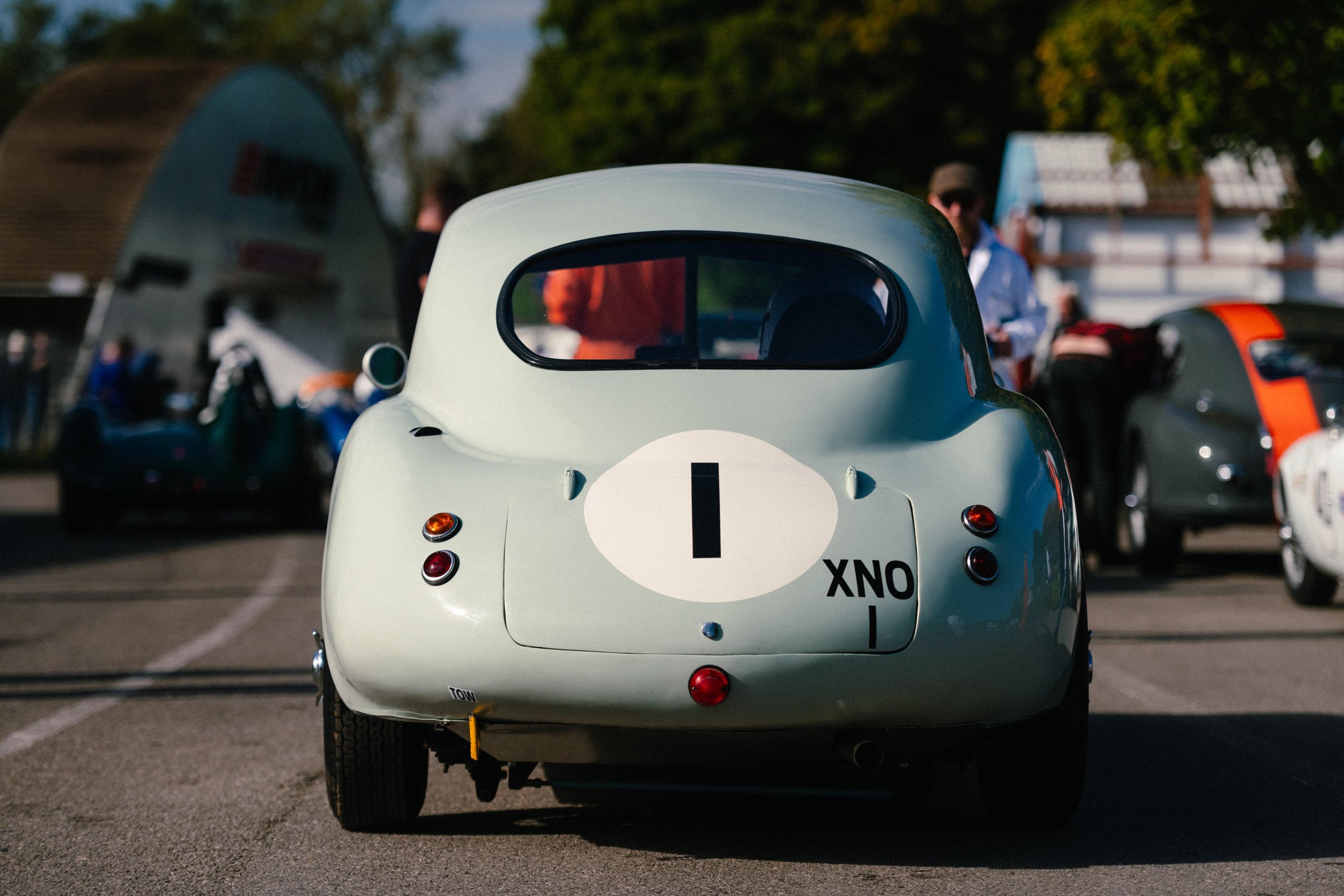 Rear view of a vintage light green race car with a white oval and black number 1 on the back, parked at a car show with other vehicles and people in the background.