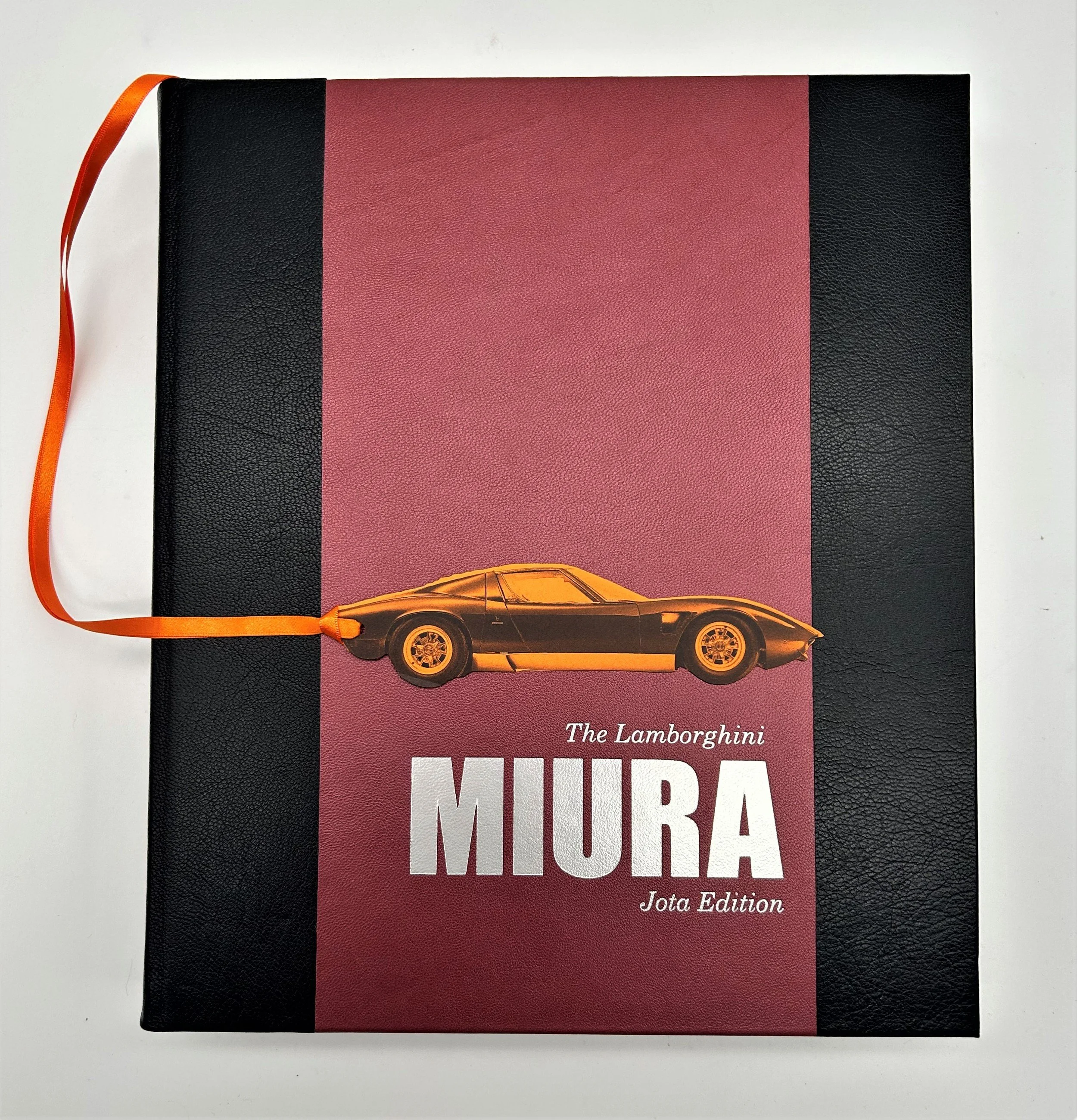 A book titled 'The Lamborghini Miura Jota Edition' with a maroon and black cover and an orange car graphic, with an orange ribbon bookmark.
