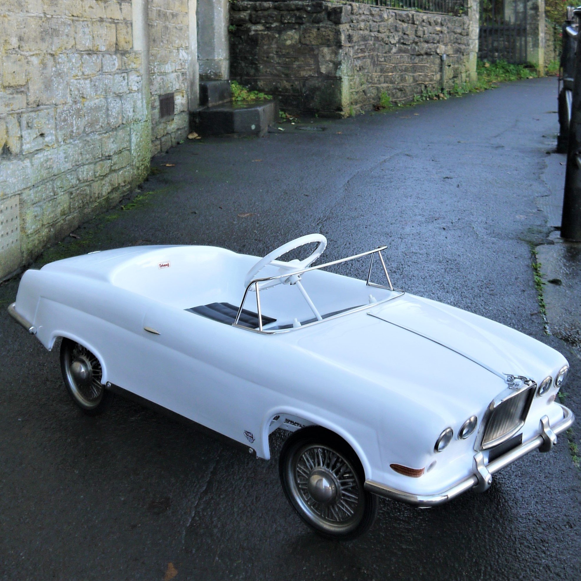 A miniature white vintage car replica parked on a wet pavement near a stone wall and black fence with a pole.