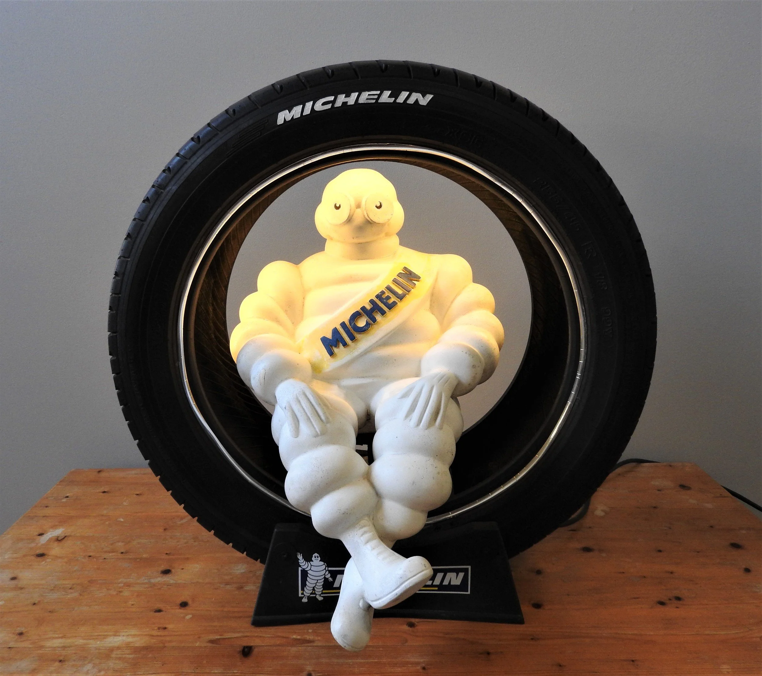 A white Michelin Man sculpture sitting with crossed legs in front of a black tire, both displaying the Michelin logo, lit by a yellow light.
