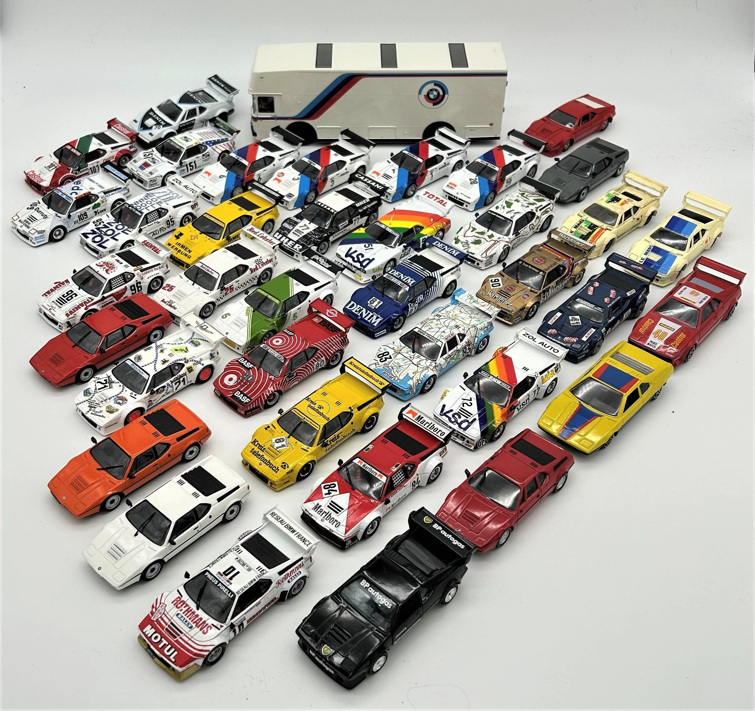 A collection of miniature race cars with various colorful designs, logos, and numbers, arranged on a white surface with a larger truck model in the background.