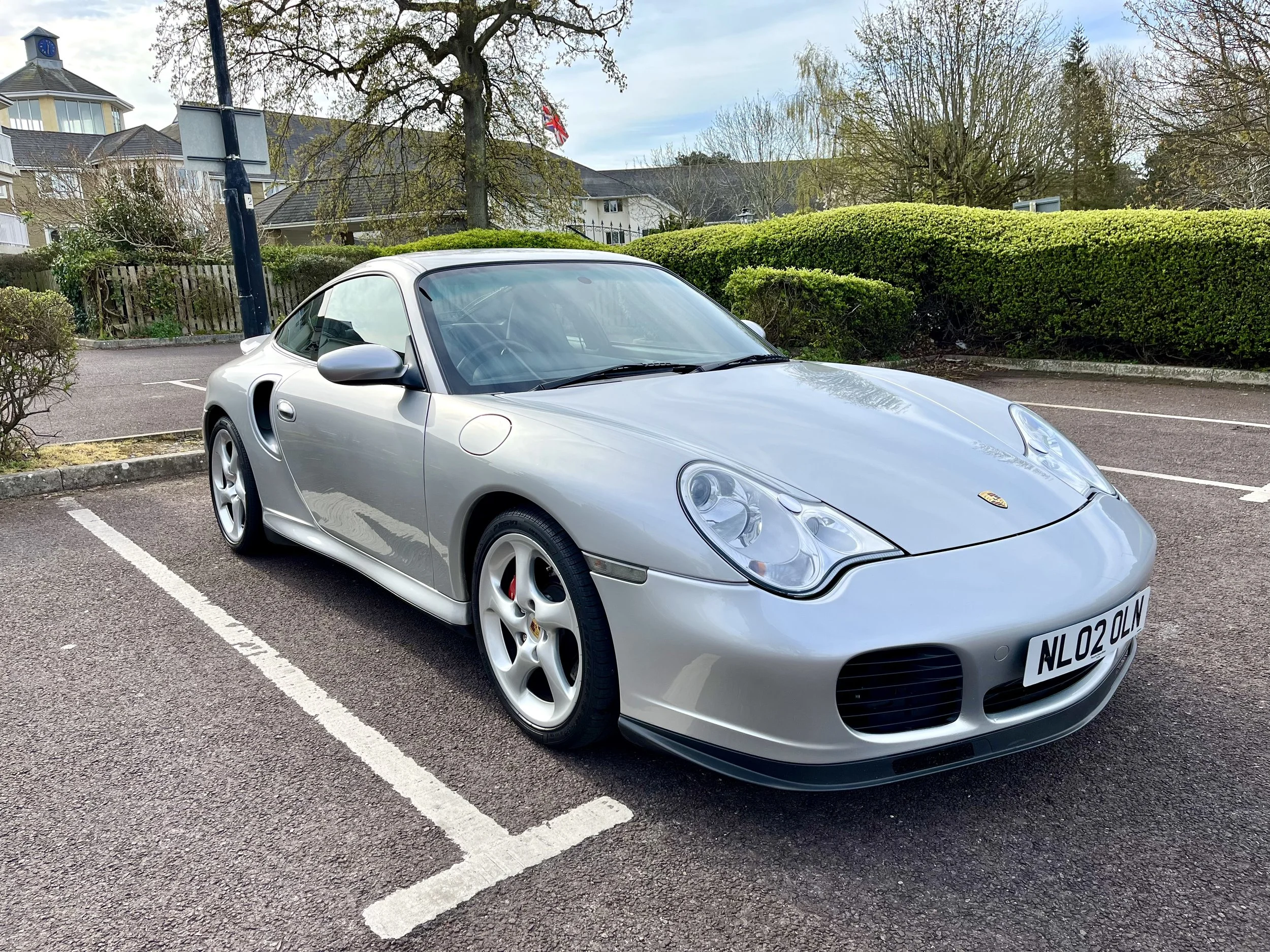 A silver Porsche sports car parked in a parking lot with green hedges and trees in the background.