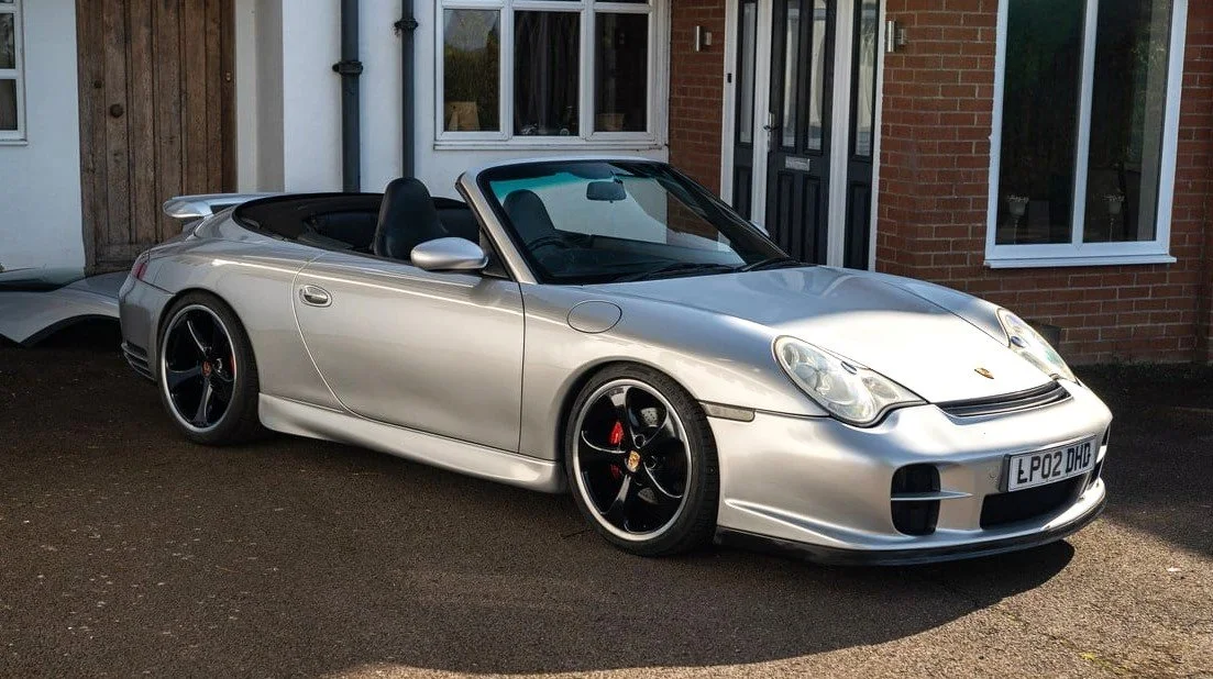 Silver Porsche convertible parked in front of a house with a brick wall and windows.