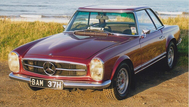 A vintage red Mercedes-Benz convertible parked on grass near a beach with ocean waves in the background.