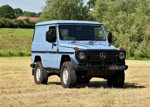 Blue Mercedes-Benz G-Class off-road vehicle parked on grassy field with trees and houses in the background.