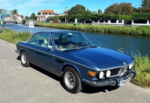 A vintage blue BMW car parked near a waterway with houses and trees in the background.