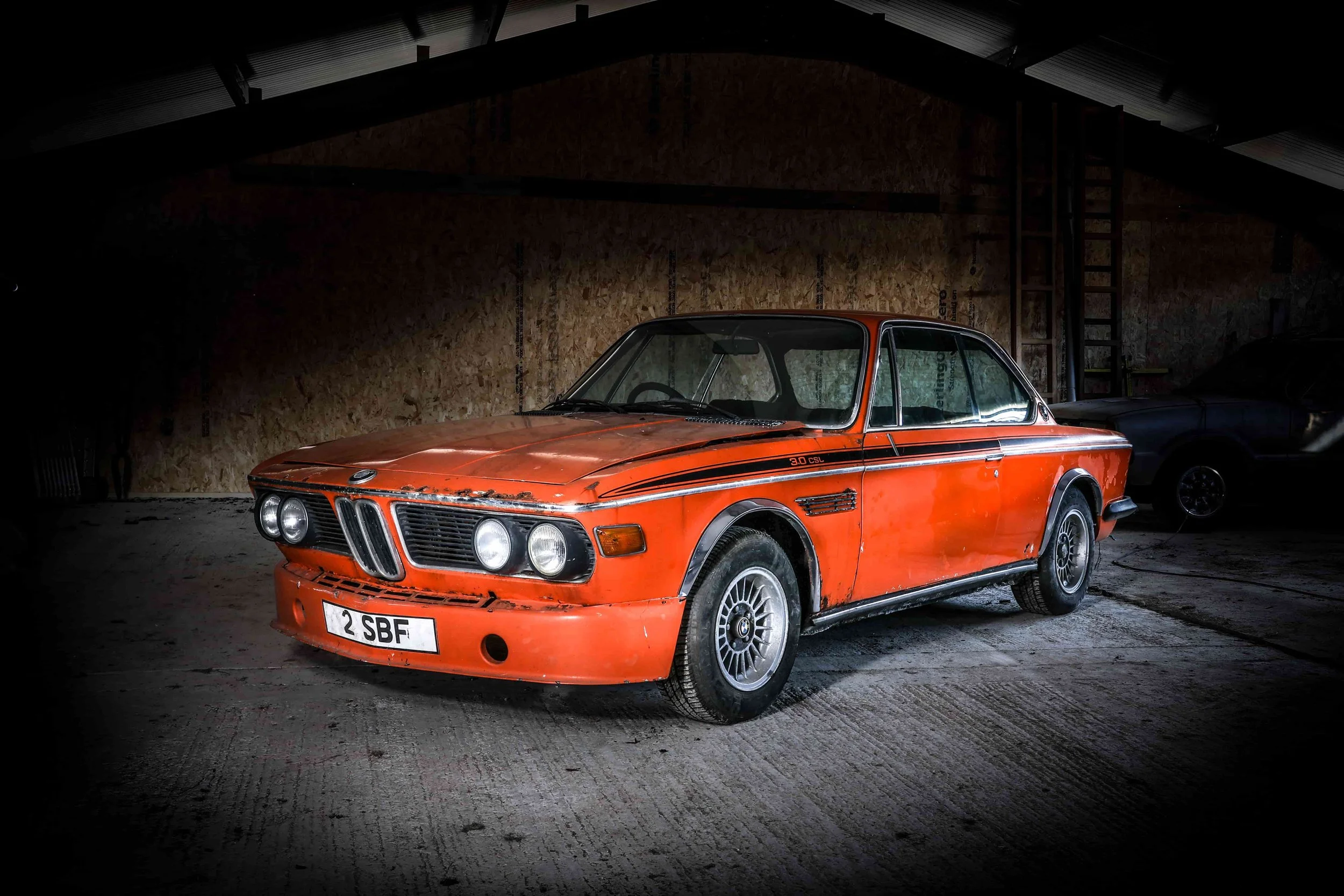A vintage orange BMW 3.0 CSL car inside a dimly lit garage or warehouse with a wooden wall background.